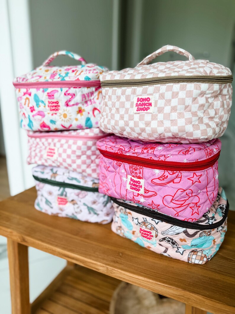 May include: A stack of quilted cosmetic bags in various patterns and colors. Designs include pink and white checkered, floral, and cowboy-themed prints. Each bag has a top handle and a zipper closure. The bags have the text "BOHO RANCH SHOP".
