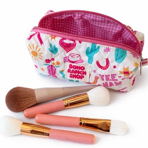 May include: A quilted white cosmetic bag with a colorful western-themed print featuring cacti, cowboy hats, and the text "BOHO RANCH SHOP" and "YEE-HAW". The bag has a pink gingham interior and is accompanied by four makeup brushes with pink handles.