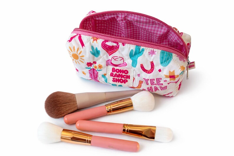 May include: A quilted white cosmetic bag with a colorful western-themed print featuring cacti, cowboy hats, and the text "BOHO RANCH SHOP" and "YEE-HAW". The bag has a pink gingham interior and is accompanied by four makeup brushes with pink handles.