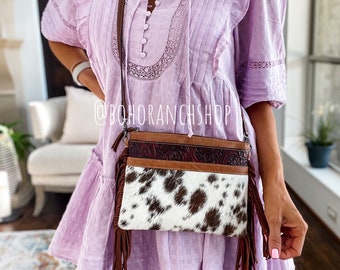 May include: A white and brown cowhide crossbody bag with brown leather trim and fringe. The bag has a floral design on the front flap and is worn over a lavender dress.