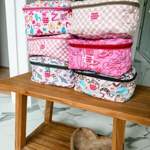 May include: A stack of quilted cosmetic bags in various patterns and colors, including pink gingham, floral, and cowboy-themed designs. Each bag has a zipper closure and a handle, with the text "BOHO RANCH SHOP" visible on several.