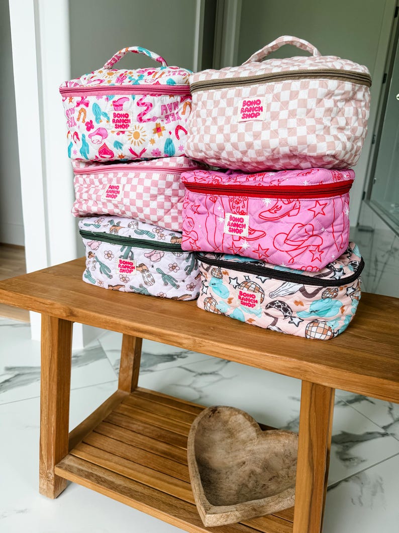 May include: A stack of quilted cosmetic bags in various patterns and colors, including pink gingham, floral, and cowboy-themed designs. Each bag has a zipper closure and a handle, with the text "BOHO RANCH SHOP" visible on several.