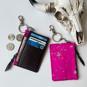 May include: Two zippered card holders with keychains. One is brown leather with a zippered pocket and the other is pink with gold splatter paint.