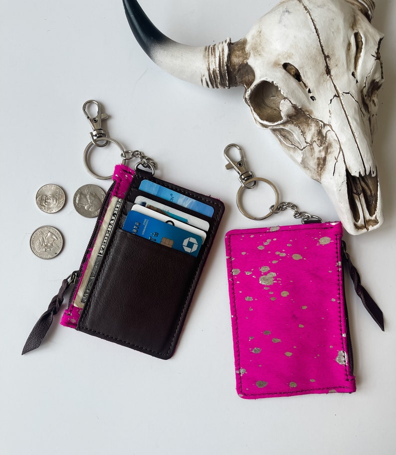 May include: Two zippered card holders with keychains. One is brown leather with a zippered pocket and the other is pink with gold splatter paint.