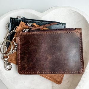 May include: Three leather wallets with zipper closures and a silver chain attached to the top wallet. The wallets are brown, tan, and black.