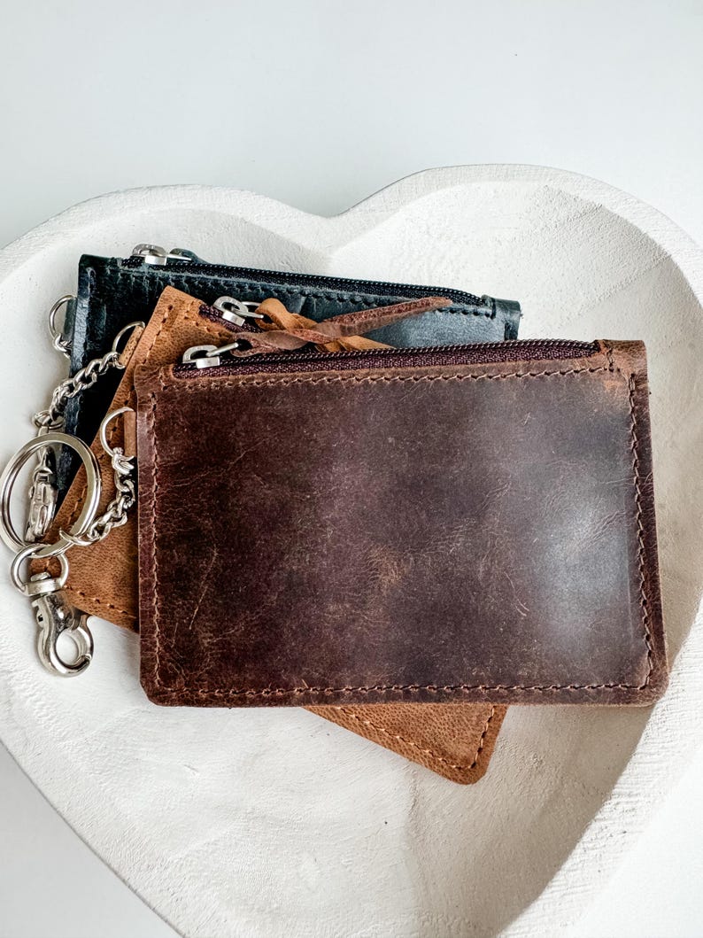 May include: Three leather wallets with zipper closures and a silver chain attached to the top wallet. The wallets are brown, tan, and black.