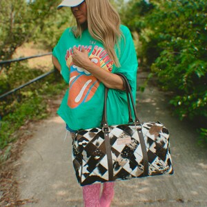 May include: A woman wearing a green t-shirt with "The Rolling Stones" printed on it, a white baseball cap with "Cowboy Hat" printed on it, and pink cowboy boots. She is carrying a black and white patchwork leather duffel bag with a brown leather handle.