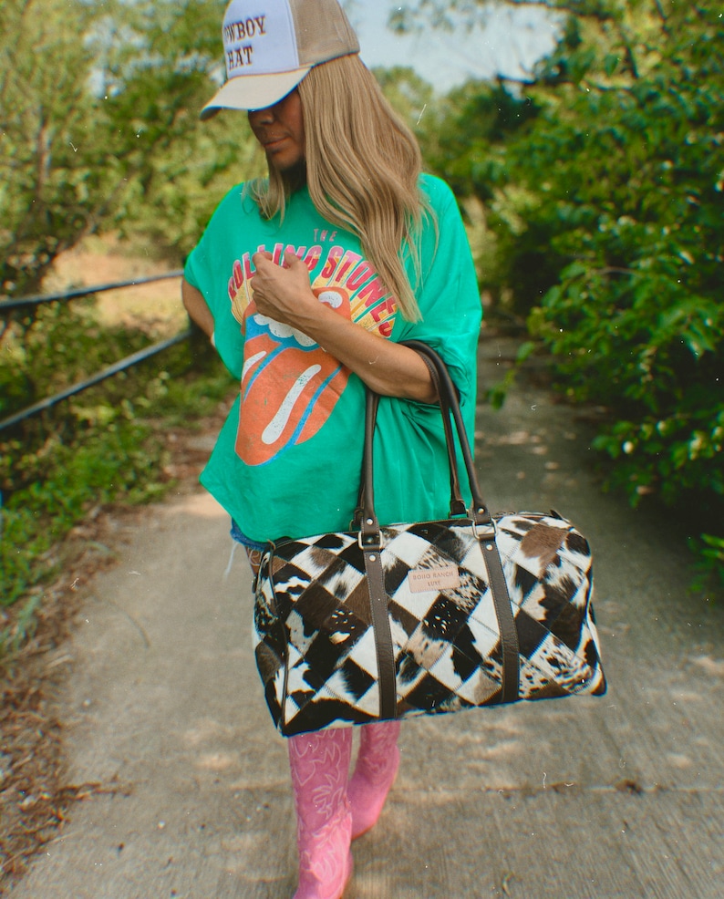 May include: A woman wearing a green t-shirt with "The Rolling Stones" printed on it, a white baseball cap with "Cowboy Hat" printed on it, and pink cowboy boots. She is carrying a black and white patchwork leather duffel bag with a brown leather handle.