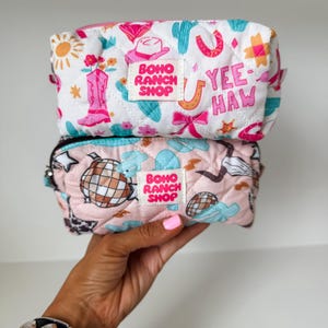 May include: Two quilted zippered pouches stacked vertically. The top white pouch features pink cowboy boots, cacti, suns, and the text YEE-HAW. The bottom pink pouch displays disco balls, cacti, and stars. Both have a BOHO RANCH SHOP label centered.