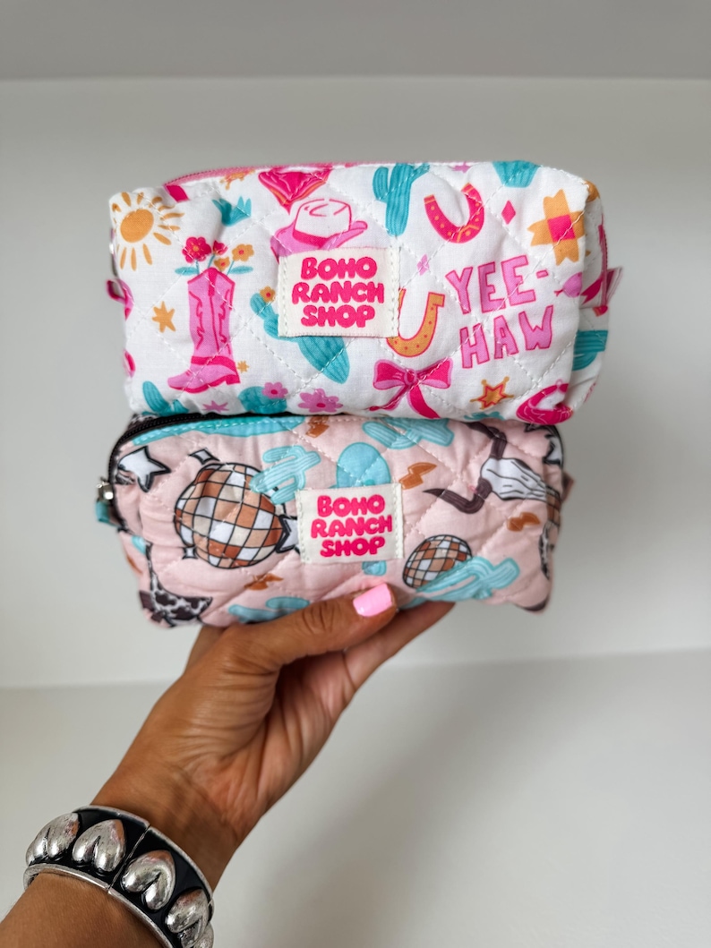 May include: Two quilted zippered pouches stacked vertically. The top white pouch features pink cowboy boots, cacti, suns, and the text YEE-HAW. The bottom pink pouch displays disco balls, cacti, and stars. Both have a BOHO RANCH SHOP label centered.