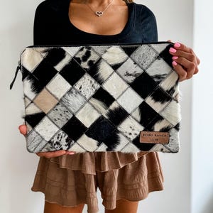 May include: A black and white patchwork cowhide clutch with a brown leather patch that says "BOHO RANCH LUXE".