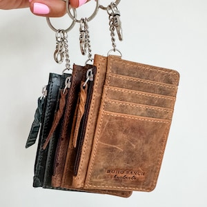 May include: Four leather card holders with zipper closures attached to a silver key chain. The card holders are brown, dark gray, and light gray. The brown card holder has the text "BOHO RANCH handcrafts" embossed on the front.