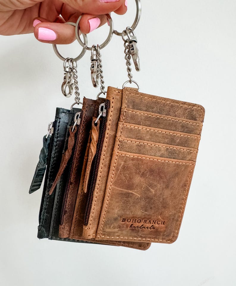 May include: Four leather card holders with zipper closures attached to a silver key chain. The card holders are brown, dark gray, and light gray. The brown card holder has the text "BOHO RANCH handcrafts" embossed on the front.
