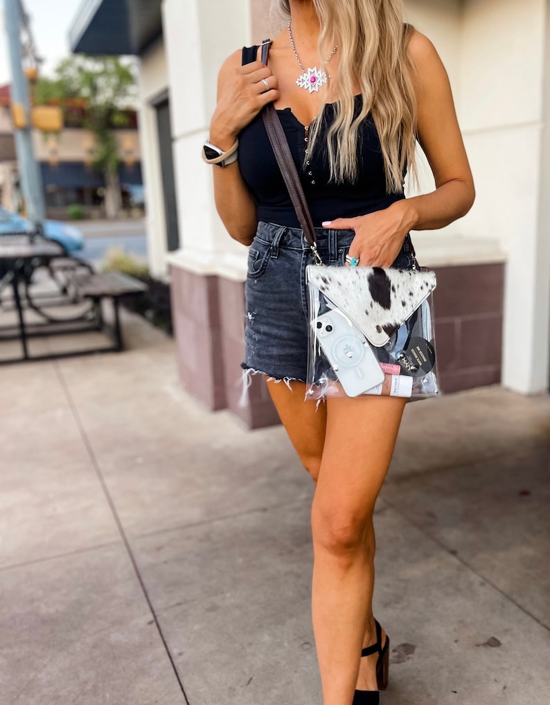 May include: A clear crossbody bag with a cowhide print flap, featuring a dark brown strap. The transparent bag contains a white smartphone with a circular ring holder, a small black circular container, and a pink tube, all visible through the plastic.