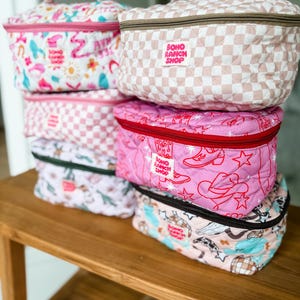 May include: A stack of quilted cosmetic bags with various patterns, including pink and white checkered, cowboy boots and stars, and a whimsical print. Each bag has a top handle and a zipper closure. The bags are labeled "Boho Ranch Shop".