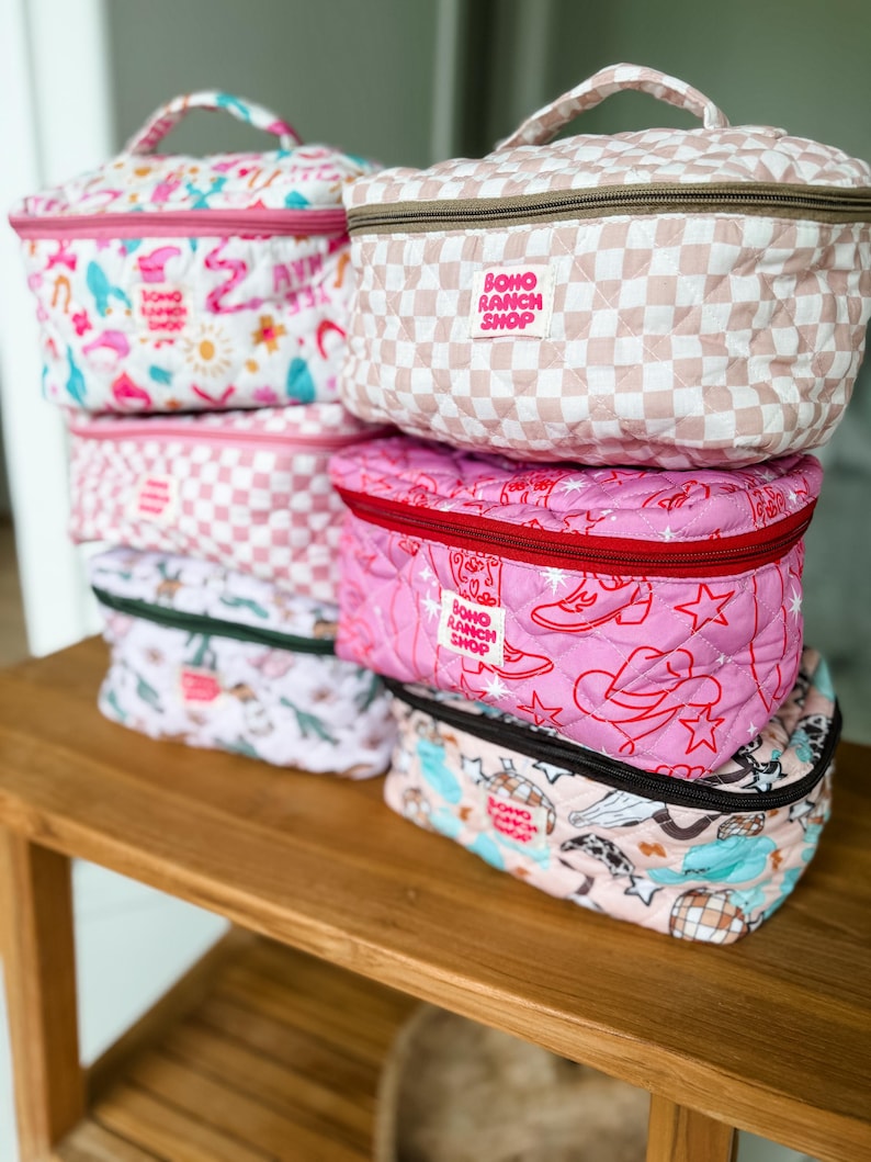 May include: A stack of quilted cosmetic bags with various patterns, including pink and white checkered, cowboy boots and stars, and a whimsical print. Each bag has a top handle and a zipper closure. The bags are labeled "Boho Ranch Shop".