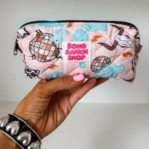 May include: A quilted light pink zippered pouch featuring a repeating pattern of disco balls, light blue cacti, lightning bolts, and cow skulls. A white rectangular label in the center displays the text BOHO RANCH SHOP in bold pink letters.