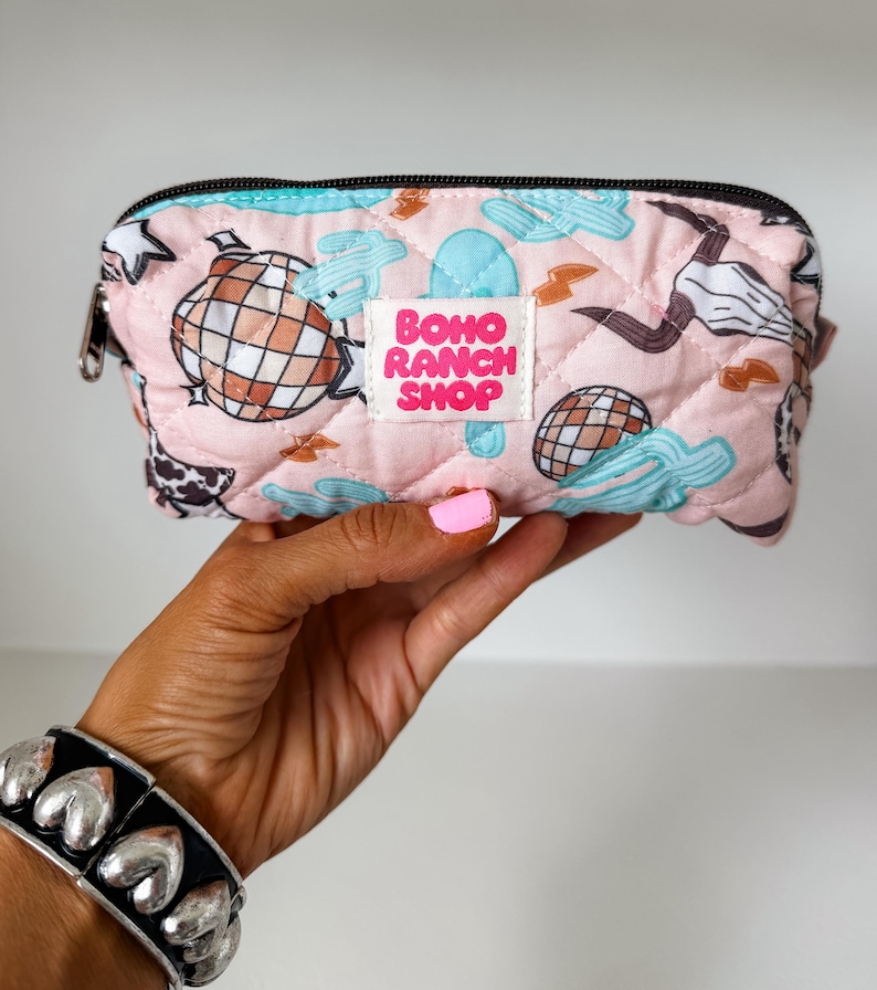 May include: A quilted light pink zippered pouch featuring a repeating pattern of disco balls, light blue cacti, lightning bolts, and cow skulls. A white rectangular label in the center displays the text BOHO RANCH SHOP in bold pink letters.