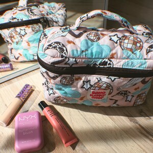 May include: Pink quilted makeup bag with a black zipper and handle. The bag features a pattern of disco balls, stars, clouds, and cow print. The text "BOHO RANCH SHOP" is visible on the bag. Makeup products are arranged around the bag.