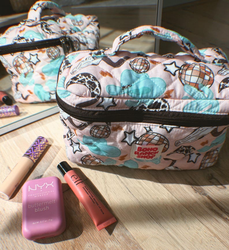 May include: Pink quilted makeup bag with a black zipper and handle. The bag features a pattern of disco balls, stars, clouds, and cow print. The text "BOHO RANCH SHOP" is visible on the bag. Makeup products are arranged around the bag.