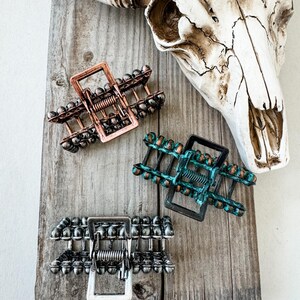 May include: Three metal hair claw clips in copper, teal, and silver tones, each with a rectangular frame and beaded accents. The clips are arranged on a weathered wooden surface, with a cow skull in the background.