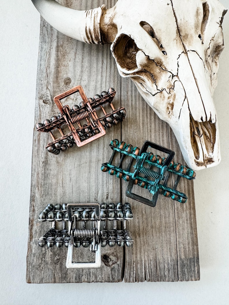 May include: Three metal hair claw clips in copper, teal, and silver tones, each with a rectangular frame and beaded accents. The clips are arranged on a weathered wooden surface, with a cow skull in the background.