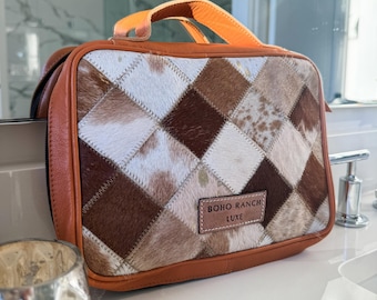 Puede incluir: Un neceser rectangular de cuero marrón con un diseño de patchwork. El patchwork presenta cuadrados de piel de animal marrón, blanco y beige. La bolsa tiene un asa y ribete de cuero marrón, y el texto "BOHO RANCH LUXE" es visible.