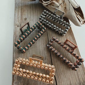 May include: Four rectangular hair claw clips in gold, silver, and copper tones, each adorned with small, round beads. The clips are arranged on a weathered wooden surface, with a cow skull in the background.