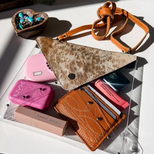 May include: A clear plastic bag containing a triangular brown and white animal print pouch, a tan leather card holder with white stitching, a pink NYX blush compact, a Fenty Beauty lipstick, and a pink Glow Reviver Melting Lip Balm tube.
