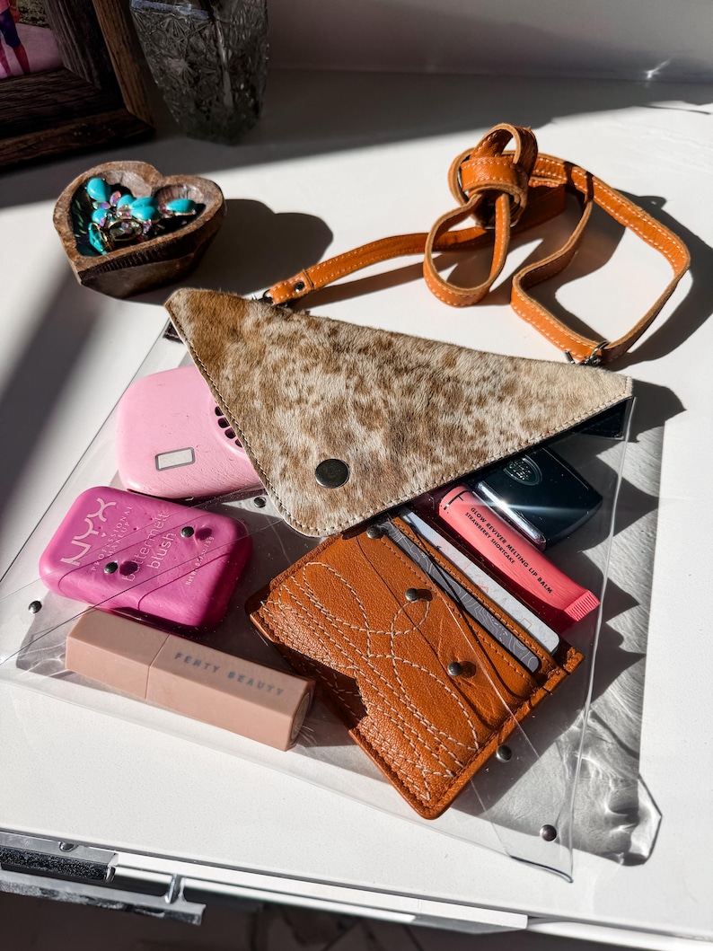 May include: A clear plastic bag containing a triangular brown and white animal print pouch, a tan leather card holder with white stitching, a pink NYX blush compact, a Fenty Beauty lipstick, and a pink Glow Reviver Melting Lip Balm tube.