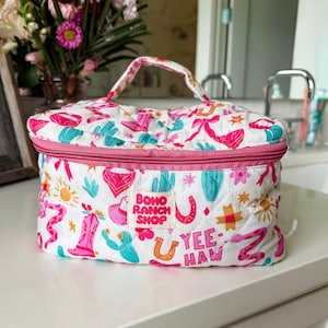 May include: A white quilted cosmetic bag with a pink zipper and handle. The bag is decorated with a repeating pattern of cacti, cowboy boots, horseshoes, and the text "Yee Haw". The label reads "Boho Ranch Shop".