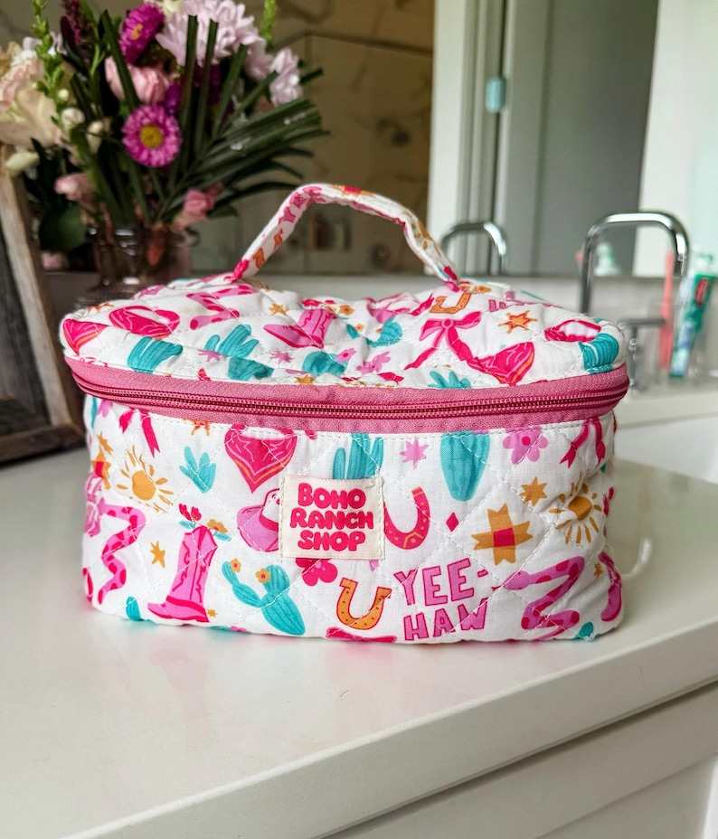 May include: A white quilted cosmetic bag with a pink zipper and handle. The bag is decorated with a repeating pattern of cacti, cowboy boots, horseshoes, and the text "Yee Haw". The label reads "Boho Ranch Shop".