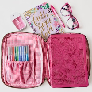 May include: An open pink gingham case with pens, a floral journal with "faith hope love" text, a pink pen, glasses, a pink floral Bible, and a pink hand sanitizer bottle. The Bible and journal have floral designs.