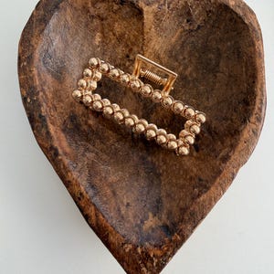 May include: A gold-colored rectangular hair claw clip with a beaded design. The clip is resting in a heart-shaped wooden bowl with a dark brown finish. The clip's spring mechanism is visible.