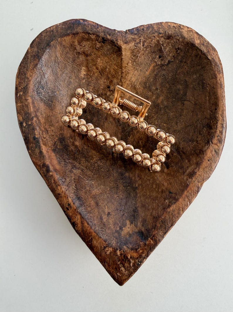 May include: A gold-colored rectangular hair claw clip with a beaded design. The clip is resting in a heart-shaped wooden bowl with a dark brown finish. The clip's spring mechanism is visible.