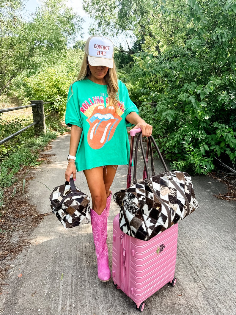 May include: A woman wearing a green t-shirt with "The Rolling Stones" printed on it, a white baseball cap that says "Cowboy Hat", pink cowboy boots, and carrying a pink suitcase and two brown and white patterned bags.