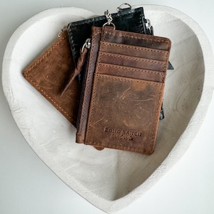 May include: Three leather wallets with a zipper closure. The brown wallet has the text "BOHO RANCH" on the front. The wallets are sitting in a white heart-shaped bowl.