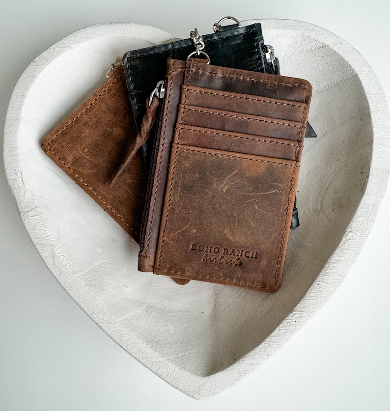 May include: Three leather wallets with a zipper closure. The brown wallet has the text "BOHO RANCH" on the front. The wallets are sitting in a white heart-shaped bowl.