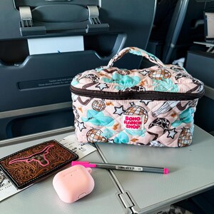 May include: A pink quilted cosmetic bag with a handle, featuring a pattern of clouds, stars, and disco balls. A brown leather wallet with a pink steer head design, pink AirPod case, and a pen are on a tray.