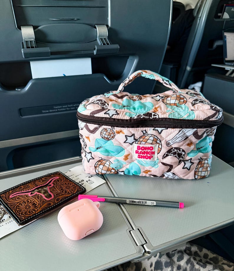 May include: A pink quilted cosmetic bag with a handle, featuring a pattern of clouds, stars, and disco balls. A brown leather wallet with a pink steer head design, pink AirPod case, and a pen are on a tray.
