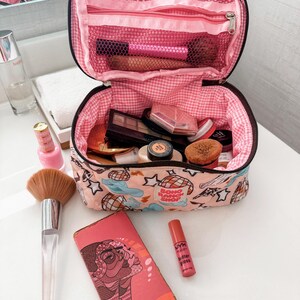 May include: Open makeup bag with pink gingham lining, filled with cosmetics. A large makeup brush, a pink compact, and a lip gloss are next to the bag. A pink patterned cosmetic bag is in the background.