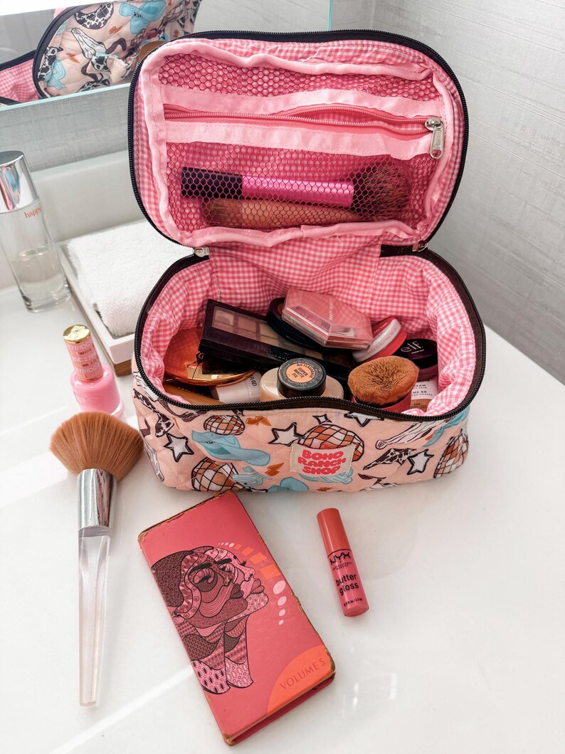 May include: Open makeup bag with pink gingham lining, filled with cosmetics. A large makeup brush, a pink compact, and a lip gloss are next to the bag. A pink patterned cosmetic bag is in the background.
