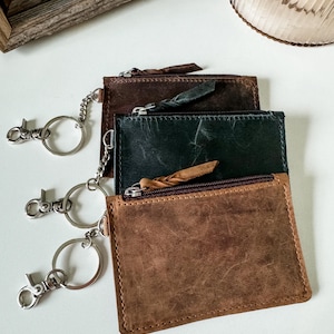 May include: Three leather keychains with zipper closures and key rings. The keychains are brown, dark green, and black.