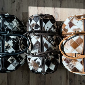 May include: Three brown, black and white patchwork leather travel bags with brown leather handles. The bags are arranged in a row on a wooden floor.