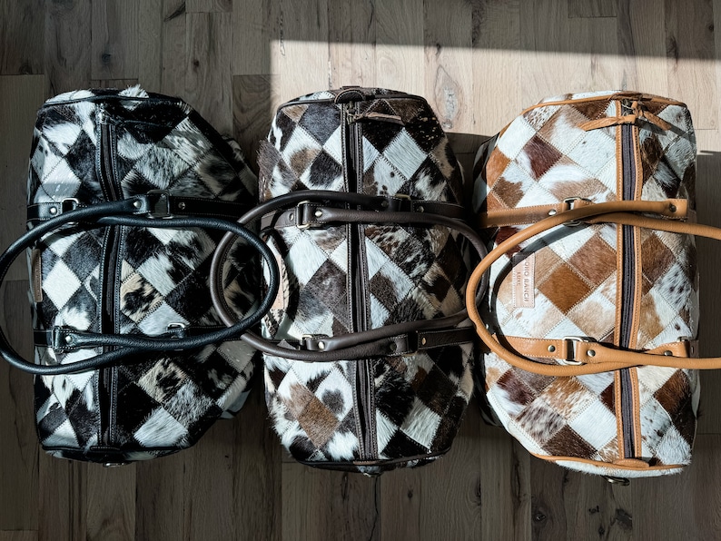 May include: Three brown, black and white patchwork leather travel bags with brown leather handles. The bags are arranged in a row on a wooden floor.