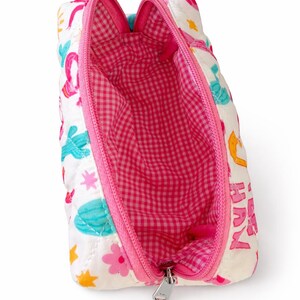 May include: A white quilted zippered pouch featuring a colorful print of cacti, flowers, and stars. The interior is lined with a pink and white gingham fabric. The pouch has a bright pink zipper closure and a silver metal zipper pull tab.