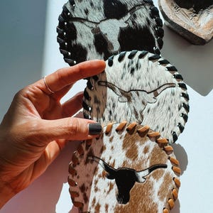 May include: Round leather coasters with a cowhide pattern and a longhorn silhouette. The coasters have a stitched edge and are in shades of black, white, and brown. A hand is touching one of the coasters.