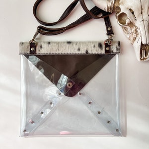May include: A clear envelope-style crossbody bag featuring a brown leather strap, a top trim of white and black cowhide, and a dark brown triangular flap with a single metal snap closure. The bag is accented with silver-toned metal studs.