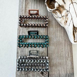 May include: Three rectangular hair claw clips in copper, teal, and silver, each adorned with a beaded design. The clips are displayed on a weathered wooden surface, with a cow skull in the background. These hair accessories offer a rustic, bohemian aesthetic.