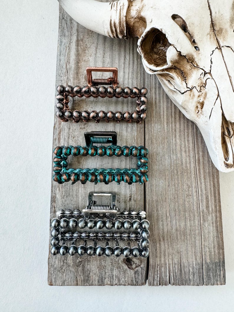 May include: Three rectangular hair claw clips in copper, teal, and silver, each adorned with a beaded design. The clips are displayed on a weathered wooden surface, with a cow skull in the background. These hair accessories offer a rustic, bohemian aesthetic.